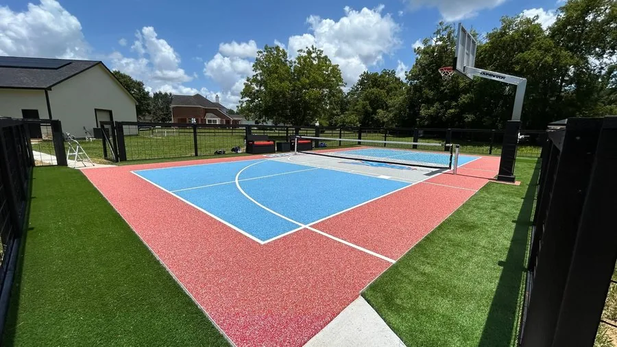 Custom blue and coral sport court with basketball hoop and pickleball net surrounded by artificial turf