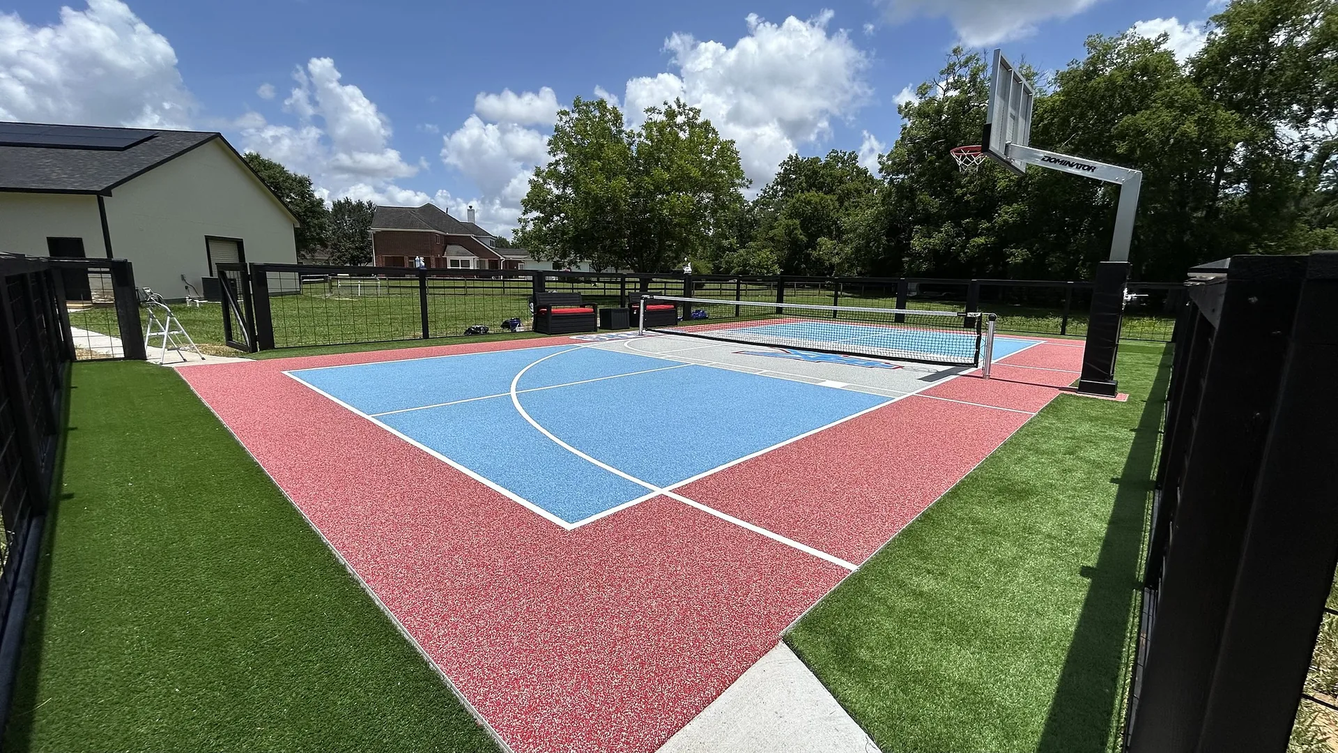 Red and blue sport court with basketball hoop and pickleball net surrounded by turf